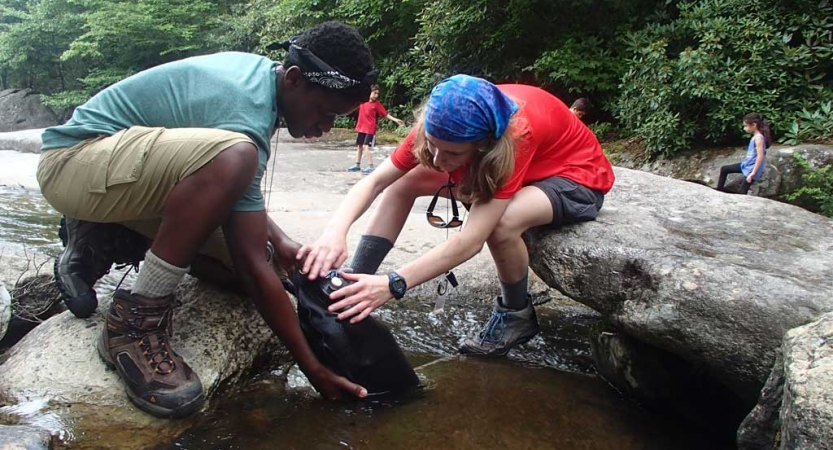 Two students kneel on rocks as they fill a water bladder in shallow water.